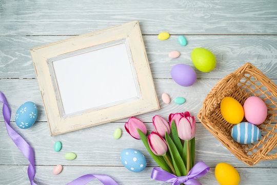 Easter holiday background with easter eggs in basket, photo frame and tulip flowers on wooden table.