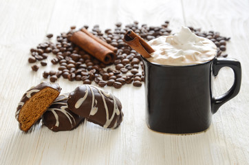 A cup of coffee with cinnamon and whipped cream. Scattered coffee beans and chocolate gingerbread on a white wooden table.
