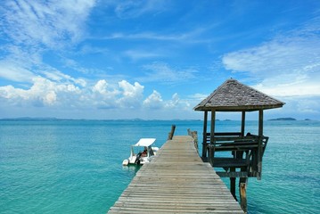 The wooden bridge stretches out to the middle of the sea with a view of the sky, and there is a white boat landing at Koh Samet Thailand.