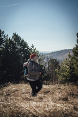 Hiking girl with backpack standing on the meadow with pine forest on the mountain and observing locality.