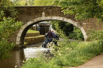 An elderly couple steer their boat under a bridge towards the canal lock gates