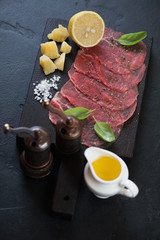 Wooden serving board with carpaccio made of raw marbled beef and seasonings, vertical shot on a black stone surface