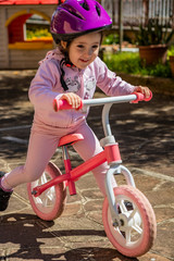 Naklejka premium A little girl dressed in pink and wearing a purple helmet plays and enjoys the balance bike. A child runs safely on a bicycle without pedals, in the backyard. Rome Italy.