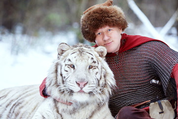 Man dressed as a medieval warrior sits in the snow in a winter forest and hugs a white tiger by the neck. The warrior is dressed in chain mail, a fur hat and a red cloak. - Powered by Adobe