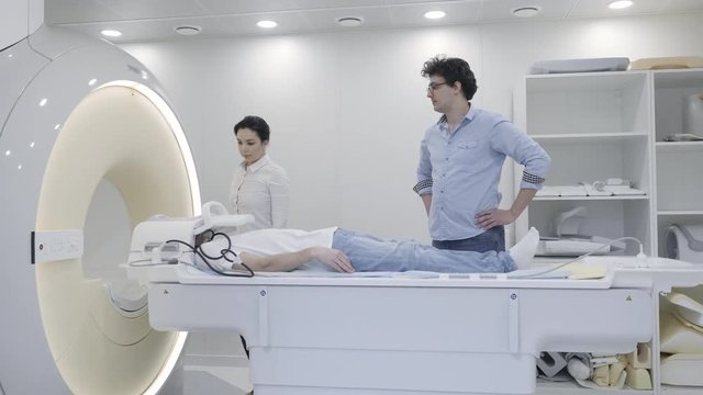 Clinic emergency MRI image head scan. Man lays in magnetic resonance imaging device during medical exam and then moving inside. Young doctor control the prosess with serious nurse together in room