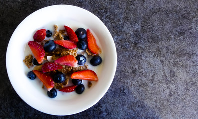Vibrant closeup of white breakfast bowl with cereal of flakes, coconut & banana, topped with blueberries and strawberries, glistening with fresh cow's milk. Dish on a black marble in natural light.