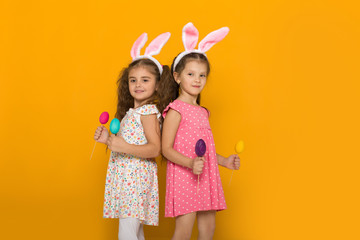 two little girls with Easter bunny ears holding colorful eggs on yellow background. Happy easter. cute children