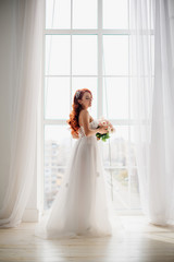 Red-haired bride in white wedding dress stands near large window