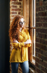 Curly redhead girl in the yellow shirt wearing spectacles standing hands crossed near the big wooden window