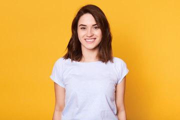 Obraz premium Closeup portrait of attractive woman stands smiling isolater over yellow background and looks directly at camera. Adorable lady wears white casual t shirt, looks happy. Copy space for advertisment.