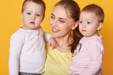 Portrait of young mother holding her little twins, cute active infants being interested to be photografed, look asides, mother looks with love on her adorable daughters. happy family concept.