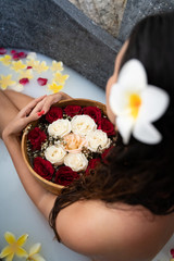Obraz na płótnie Canvas Young woman relaxing in black stone bath with tropical flowers and rose petals. Skin treatment, luxury spa concept.
