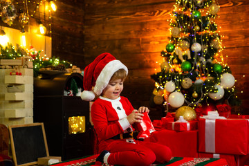 Obraz na płótnie Canvas Christmas Kids. Happy little kid is wearing Santa clothes, playing with Christmas gift box. Fireplace background. Christmas presents.
