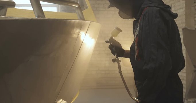 In slow motion a professional man (guy) in a garage (service) in working clothes dressed in a sweater sets up a paint gun (spray gun) and starts painting (putting a layer of ceramics) a yacht.