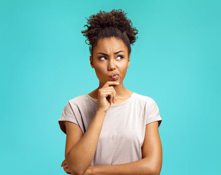 Pensive girl being deep in thoughts, raises eyebrows, curves lips, holds chin. Photo of african american girl wears casual outfit on turquoise background. Emotions and pleasant feelings concept.