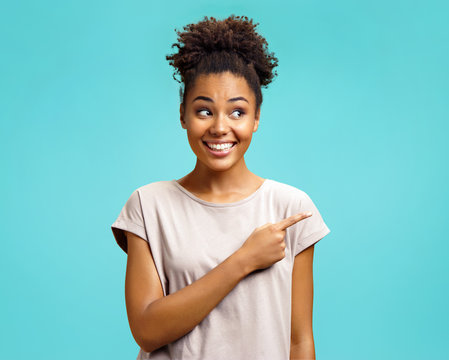 Positive girl indicates with forefinger, shows place for your advertisement. Photo of african american girl wears casual outfit on turquoise background. Emotions and pleasant feelings concept.