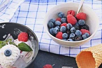 A dark bowl full of wild berry ice cream balls with mint leaves and ice cream waffles next to it on a dark kitchen surface - concept with refreshing fruit ice for the summer