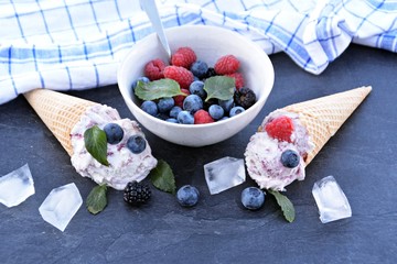 A dark bowl full of wild berry ice cream balls with mint leaves and ice cream waffles next to it on a dark kitchen surface - concept with refreshing fruit ice for the summer