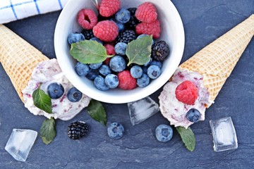 A dark bowl full of wild berry ice cream balls with mint leaves and ice cream waffles next to it on a dark kitchen surface - concept with refreshing fruit ice for the summer