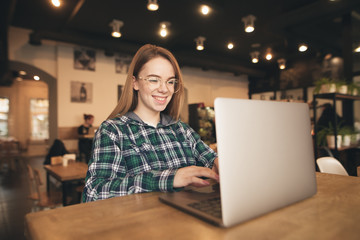 Portrait of a happy freelancer sits in a cozy cafe with a laptop, dials text on the keyboard and smiles. Positive blogger girl works in a cafe with a laptop. Student studying in a cafe.