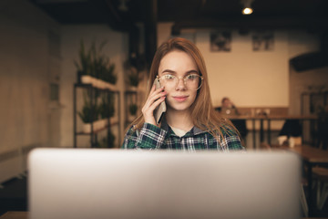 Portrait of a beautiful girl in casual clothes and glasses talking on the phone and using a laptop in a cozy cafe. Student girl works in a cafe.