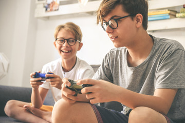 Young brothers enjoy video game at home. Smiling kids playing with console sitting on a sofa Teenager and brother playing with wireless remote controller Wifi technology is everywhere Friends have fun