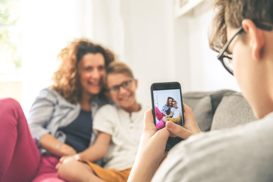 Young boy takes picture of his mother and son sitting on a sofa at home. Smiling family play with smartphone Mum and son enjoy a day off with older brother Familiar scene. Focus on cell phone