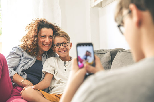 Young boy takes picture of his mother and son sitting on a sofa at home. Smiling family play with smartphone Mum and son enjoy a day off with older brother Familiar scene of happy people taking selfie