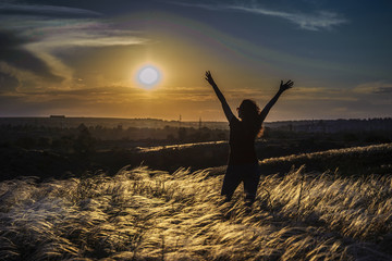 Girl in excited stands with raised hands in a feather grass on the background of the sunrise
