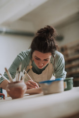 Charming young woman in apron painting pottery at workshop