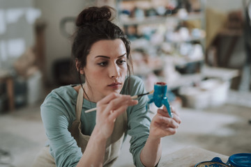 Beautiful young woman painting candleholder in blue color at workshop