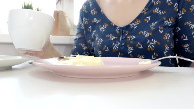 Woman sitting by the table, eating an omlet with a folk and a knife and drinking coffee or tea for breakfast at home or restaurant