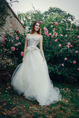 Young beautiful model is posing in a long ivory dress in the garden with a crown on her head 