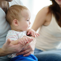Loving parents sitting in living room at home affectionately cradling adorable baby son in their arms looking at him. Childhood, parenthood, togetherness, love, happiness concept