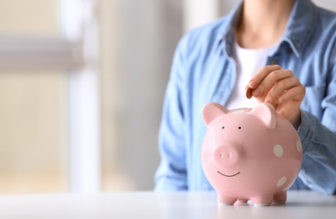 Woman putting coin into piggy bank at table indoors, closeup. Space for text