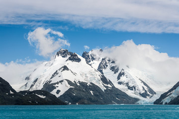 Fototapeta na wymiar Snow and glacier ice covered rocky mountain peaks, with low white clouds, glacier melt blue water, and blue sky and white clouds above, Drygalski Fjord, South Georgia