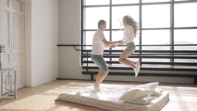 Beloved are jumping and hugging in bed holding hands in their new flat. Side view. Relationship concept.