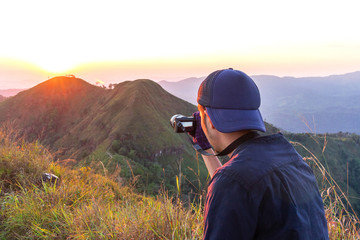 Thai young hiker taking photo of amazing sunset landscape on peak mountain at Khao Chang Puak national park in Kanchanaburi province, Thailand.