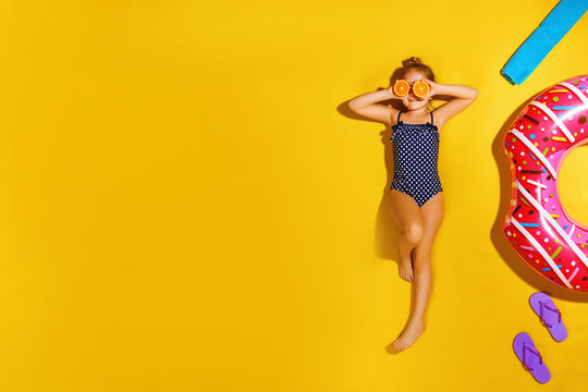 Little girl in a bathing suit lying on a donut inflatable circle. A child holds halves of an orange instead of eyes. Yellow background. View from above. Summer concept.