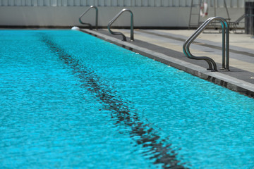 A swimming pool with a sunny reflection and beautiful wavy water surface in turquoise blue color with grab bars ladder on the sides and black line lanes underwater.