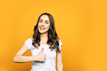 Obraz premium Tip top. “Cool” sign made by glad pretty girl with brilliant white smile, curly thick hair and hazel eyes in casual outfit in front of warm orange color.