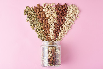 Different types of nuts and seeds in a glass jar on pink background, top view