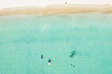 Stunning aerial view of two unidentified people doing stand up paddle board and windsurf on a clear and turquoise sea while a woman sunbathing on a white beach. Surin beach, Thailand.