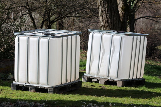 Brand new two intermediate bulk containers or IBC plastic tanks with metal cage used for water storage put on wooden pallets in local garden surrounded with grass and trees on warm sunny spring day