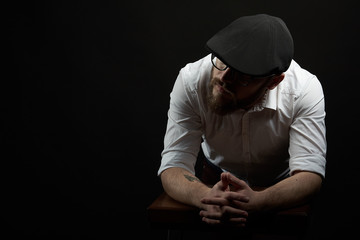 young handsome man in the glasses of struggle and mustache in a black cap and a white shirt conceived bent on his hands, a dark studio background