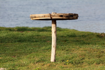 Homemade round wooden table mounted on single wooden pole in middle of uncut grass of local river bank with river in background on warm sunny spring day
