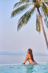 Girl in a swimsuit in the pool looks into the distance on the ocean in the background palm tree