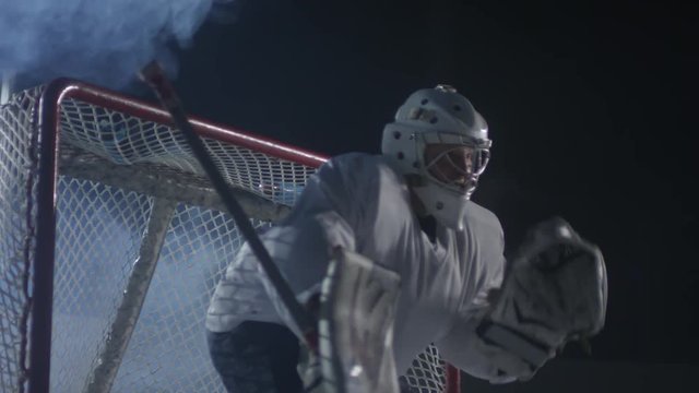 Medium shot of ice hockey goaltender in helmet standing in front of the net and catching puck with glove
