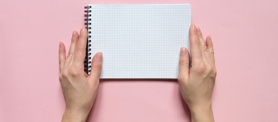 Female hands holding a notebook with blank sheets for writing on a pink background. Flat lay top view.