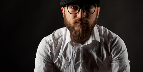 Portrait of a young man in glasses with a beard and a mustache in a black cap and a white shirt, A...